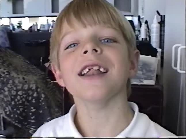 Smiling blond boy in a white polo talks playfully to the camera inside a busy hair salon