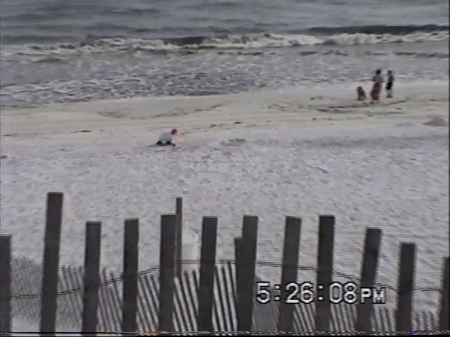 Vintage VHS footage pans from the iconic Seaside, Florida obelisk and beach houses to the ocean where people play in the sand