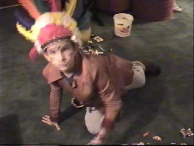 A boy dressed in a Native American Halloween costume kneels on the floor surrounded by his candy in a vintage home video