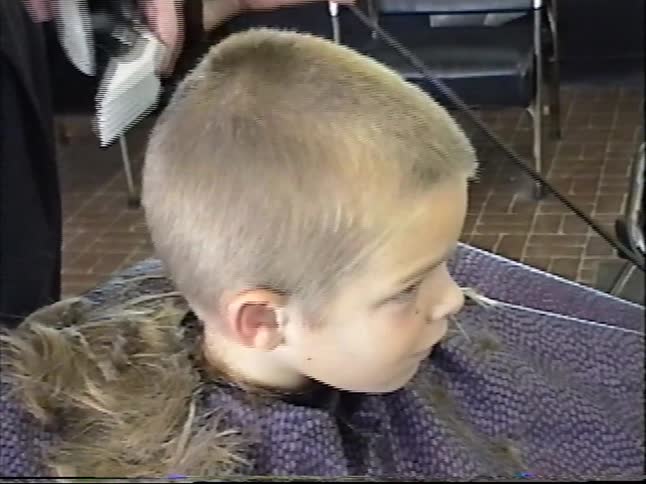 A hairstylist uses electric clippers to give a young boy a short buzz haircut in a salon