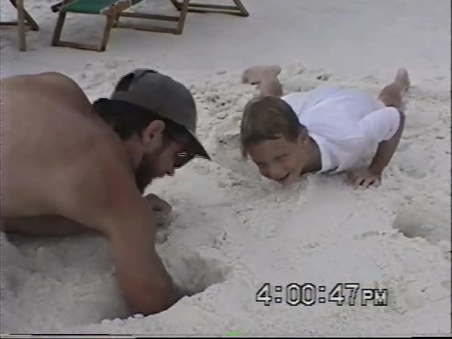 A father and son have fun digging in the sand together on a busy beach in this vintage home movie footage