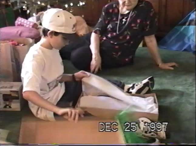 Vintage VHS footage from 1997 shows a young boy unwrapping a Christmas present as his grandmother watches by the tree