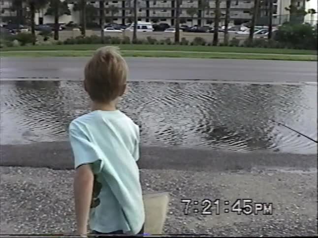 A vintage home video shows two young boys playfully pretending to fish with a rod and net in a large puddle in a parking lot