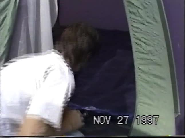 A bearded man inflates an air mattress with an electric pump inside a camping tent in the woods in 1997