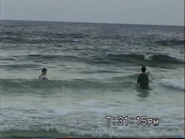 Vintage VHS footage shows two boys playing in the ocean waves in slow motion as the camera pans out to reveal the wider beach scene