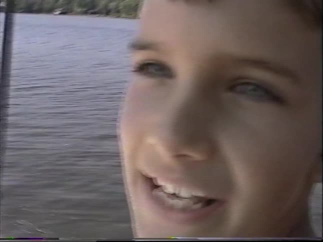 Teen rides an inflatable tube behind a speeding boat across a calm lake with a wooded shoreline