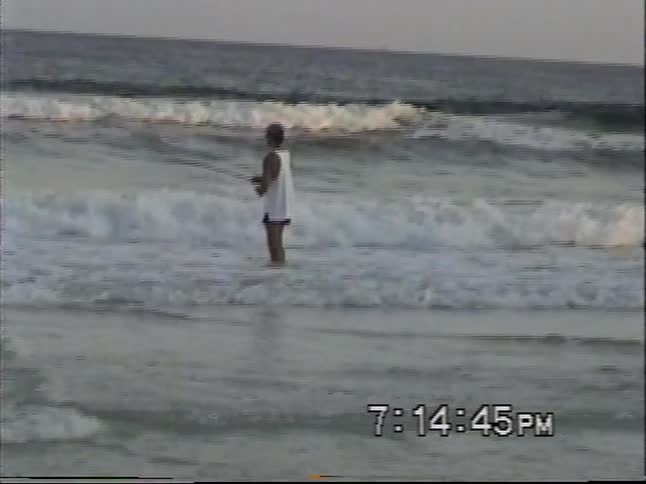 Vintage home video captures two young boys playing in the sand and a man fishing in the surf at sunset