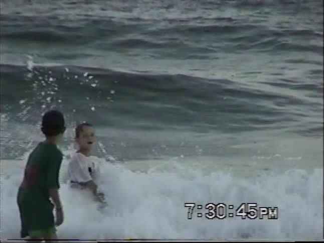 Two young boys play in the crashing ocean waves in this slow-motion vintage home video