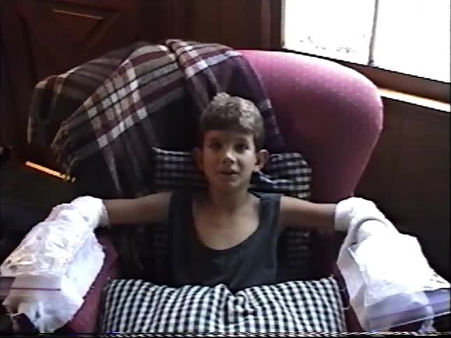 A young boy with bandaged hands covered in ice packs talks to the camera while resting in a chair