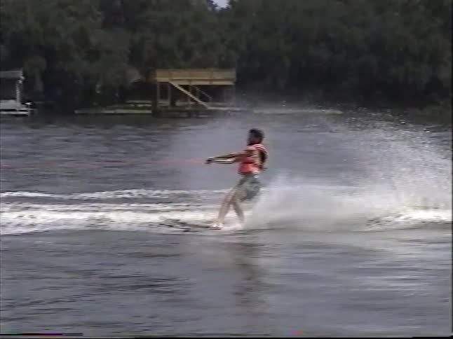 Vintage VHS footage shows a man slalom water skiing on a calm lake, waving at the camera before skiing into the distance