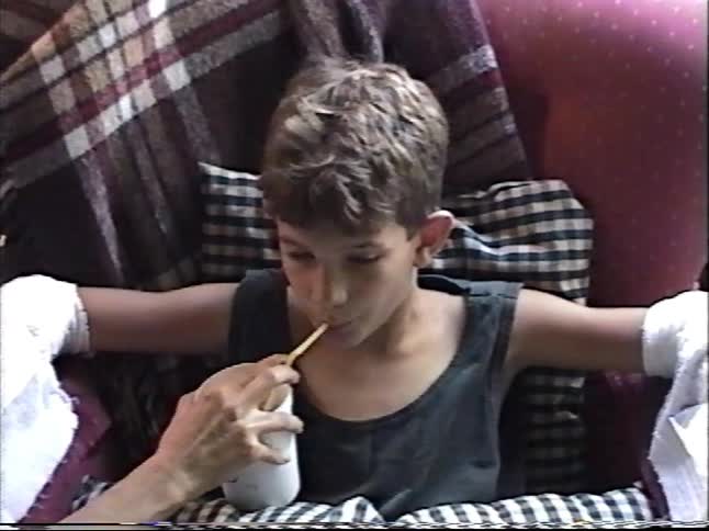 An injured boy with ice packs on both arms is helped to drink from a straw while recovering on a comfy chair at home