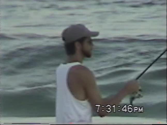 Vintage VHS footage shows a man standing in the ocean surf as he reels in his fishing line while waves crash around him