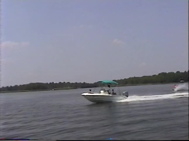 Vintage footage shows a person water skiing behind a motorboat with a family on a wide lake during a sunny day
