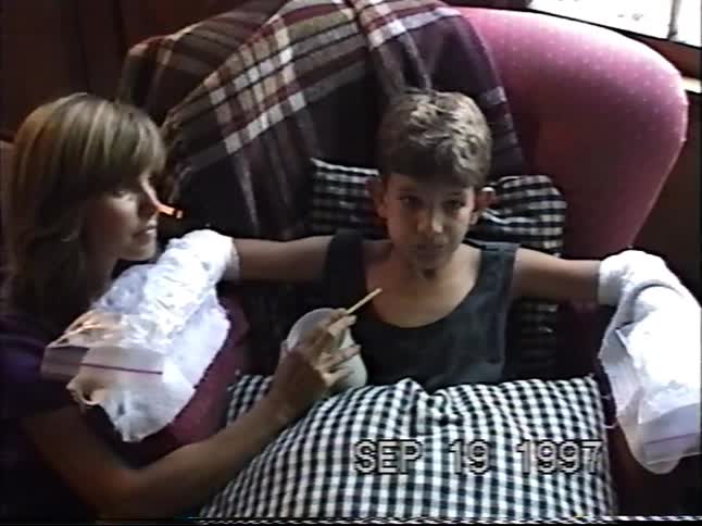 A mother cares for her son, who sits in a chair with ice packs on his arms while drinking from a cup in 1997