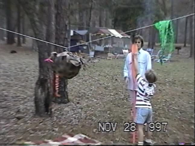 A blindfolded child hilariously swings a giant oar at a turkey-shaped piñata during a family Thanksgiving celebration in 1997