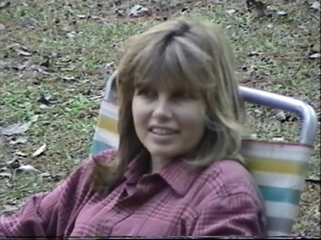 A woman relaxes in a camp chair while children gather around a campfire during a nostalgic outdoor trip in the woods