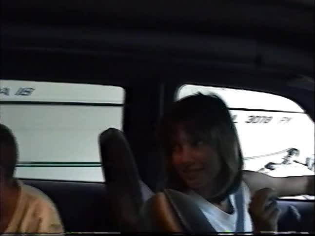 A woman chats with two boys in the back seat as they drive past boats in storage, captured on vintage VHS footage