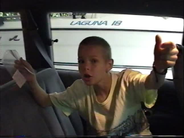 A young boy with a buzz cut excitedly gives a thumbs up from the backseat of a car in a vintage home movie
