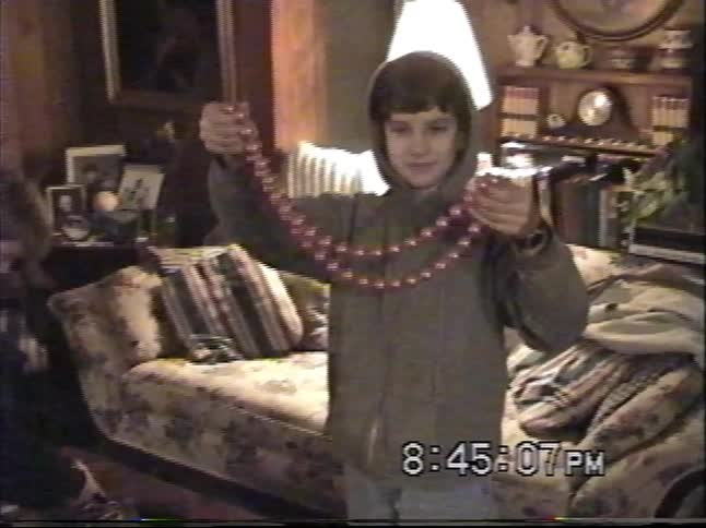 A boy in a hooded sweatshirt plays with a red beaded necklace in a living room, captured on nostalgic VHS footage