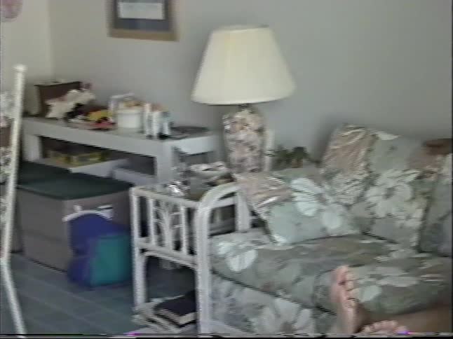 Vintage VHS footage pans from a CRT television to a man relaxing on a floral couch and making peace signs for the camera