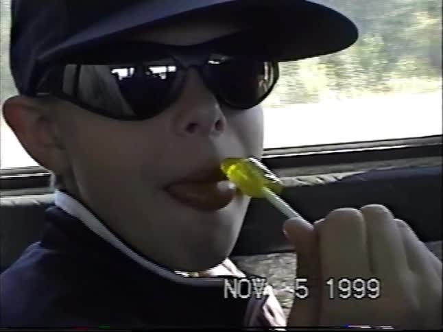 A cool kid in sunglasses and a hat enjoys a lollipop during a car ride in 1999, smiling and showing off his blue tongue