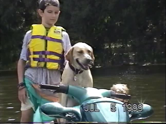 A vintage home movie from 1999 shows two young boys and their yellow lab posing on a jet ski on a lake on a summer day