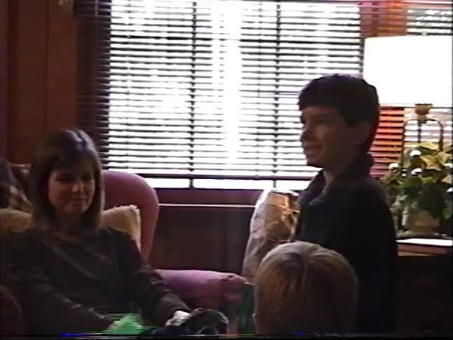 A mother and her two young sons enjoy opening presents next to the Christmas tree in a cozy living room on a vintage home video
