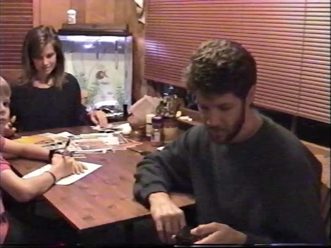 A candid 1990s home video captures a family sitting around a dining table while the father talks to the camera