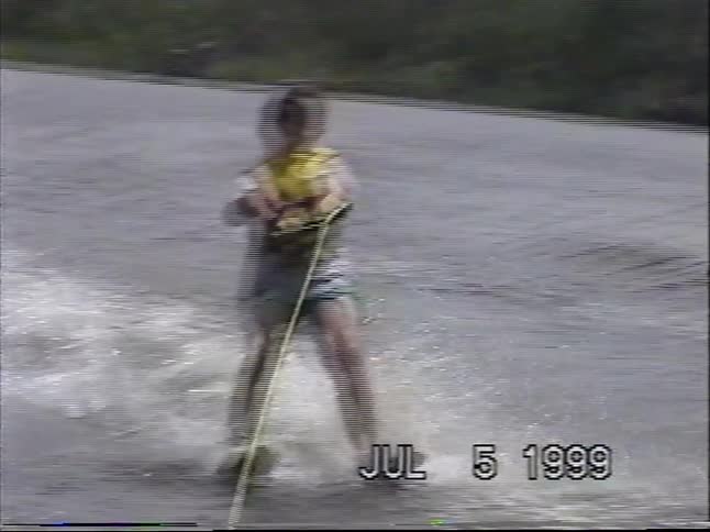 Vintage 1999 VHS footage captures a young person skillfully water skiing across a lake on a summer day