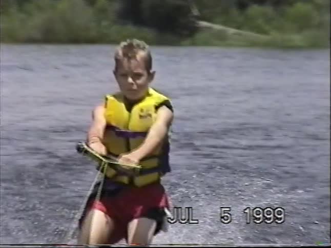 A young boy wearing a life vest successfully water skis across a lake on a summer day in this vintage home movie from 1999