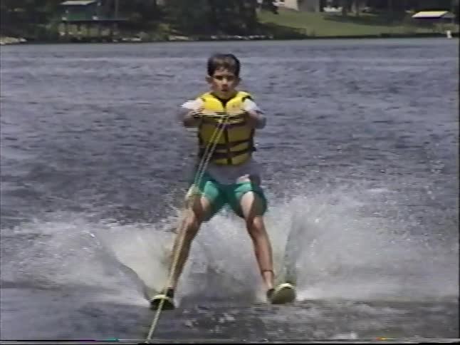 Vintage home movie footage shows a young boy in a life vest smiling as he water skis across a lake