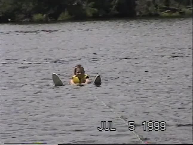 A young boy in a life vest floats in the lake with his skis on, getting ready to be pulled by the boat in a 1999 home video