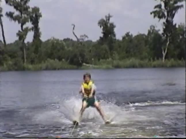 Vintage home video of a young boy learning to water ski, getting up on his skis and then falling into the lake