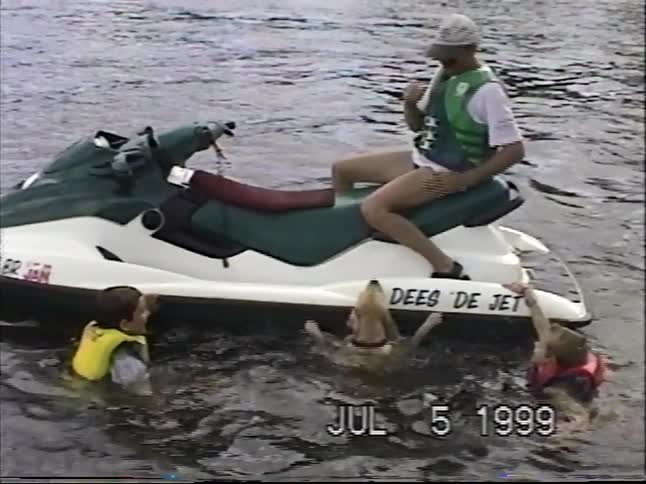 Vintage 1999 footage of a man and his dog on a jet ski, where the dog jumps in the lake to play with two swimming children