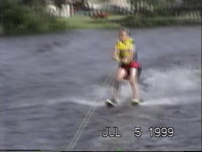 A vintage home video from 1999 captures a young boy confidently water skiing on a lake before eventually taking a splash