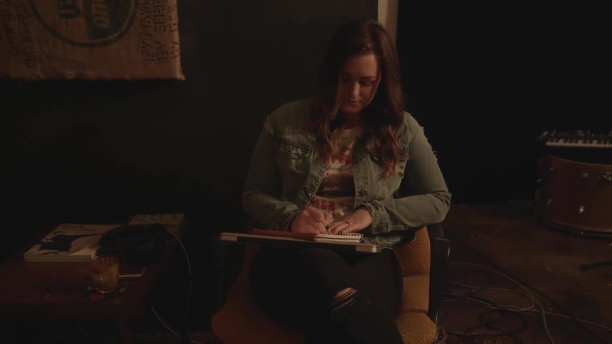 A woman is sitting in a music studio writing on a pad of paper