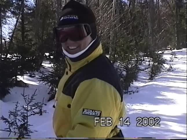 A man in a yellow ski jacket and goggles smiles for the camera while standing in a snowy forest in this vintage home video