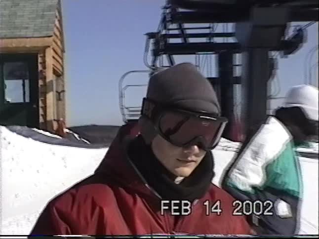 Vintage 2002 footage of a young person in full ski gear standing near a chairlift on a sunny day