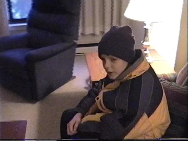Vintage home video footage zooms in on a young boy in his winter coat and beanie sitting inside on a couch