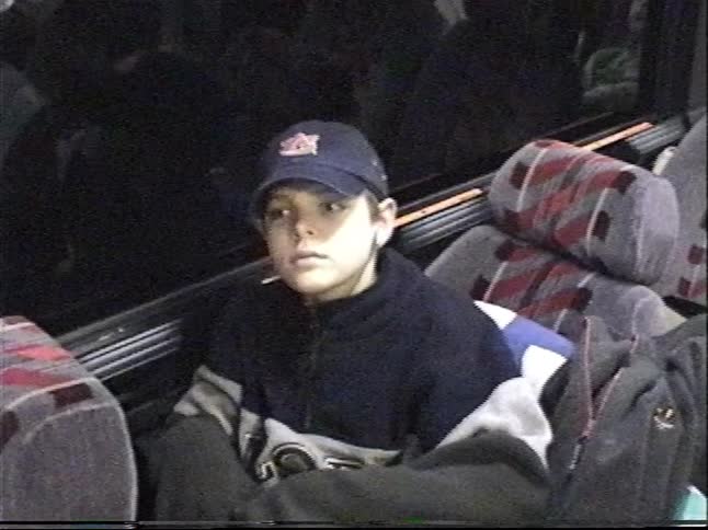 A pensive young boy wearing a baseball cap sits by the window on a moving bus at night
