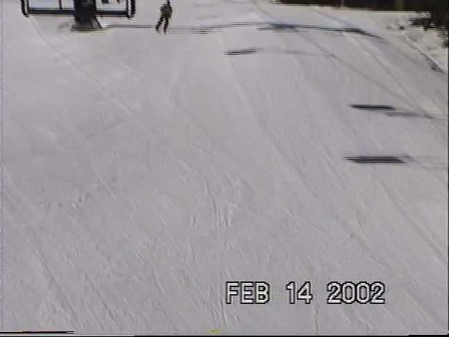 A vintage point-of-view shot from a ski lift follows a lone skier down a snowy mountain slope on a sunny day