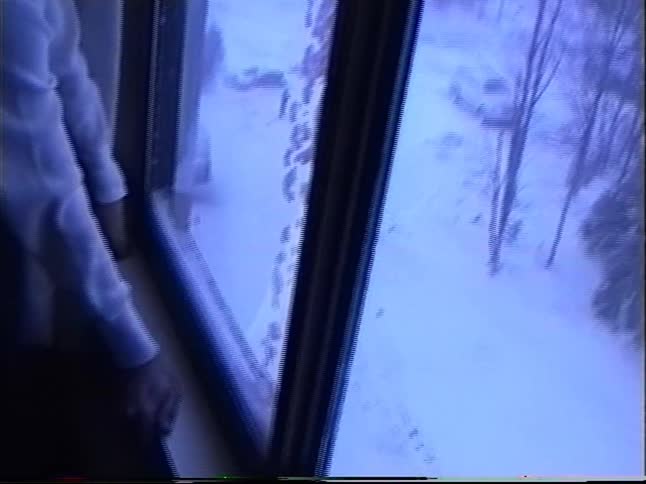 A young boy wistfully looks out a large window at a snowy winter landscape and parking lot below