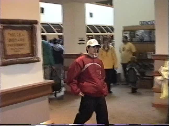 A young boy wearing a red jacket and baseball cap walks through a busy community building in this vintage home video