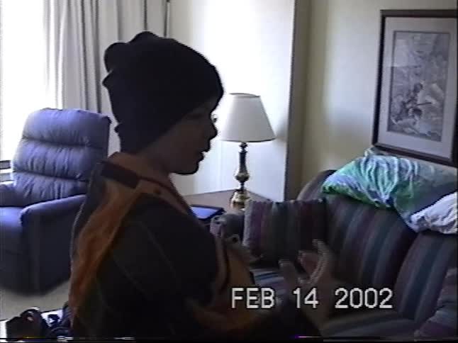 Vintage home video footage from 2002 captures a young boy excitedly putting on his jacket and backpack before giving a shaka sign to the camera