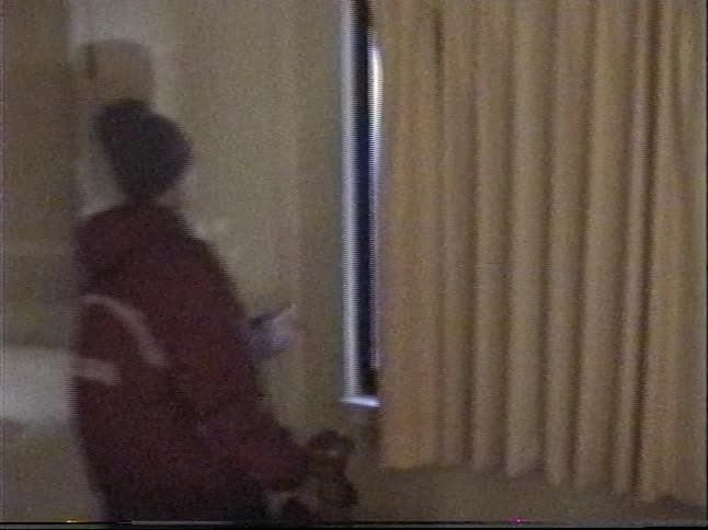 Vintage home video footage pans across a hotel bedroom, stopping on a person in winter gear who looks out the window