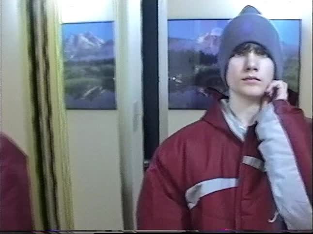 A vintage home movie clip shows a teenage boy in a red puffer jacket and beanie getting ready to go out