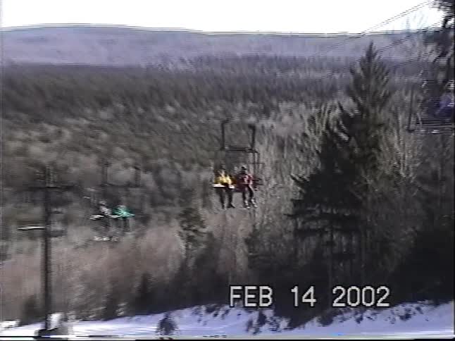 Skiers ride a chairlift up a mountain slope on a sunny winter day in this vintage 2002 home movie