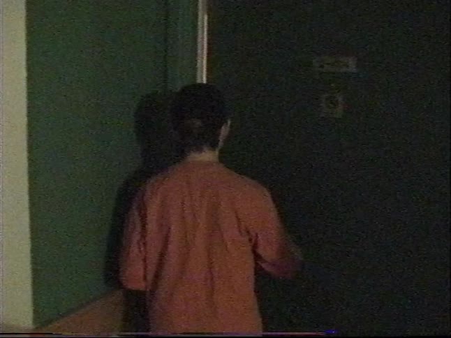 Vintage home movie footage shows two young men walking down a long, dimly lit hallway to a door