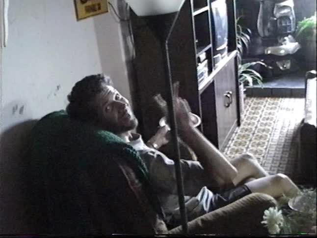 A man with a beard gestures expressively with his hands and legs while eating and talking from a recliner chair in a living room