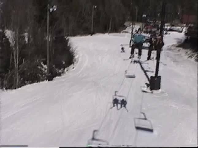 A vintage POV from a ski lift follows an adaptive skier down the snowy mountain slope on a sunny winter day